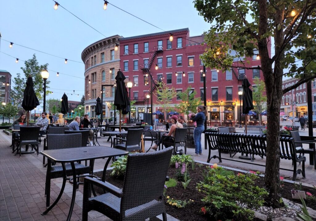 Outdoor dining area with tables, chairs, and string lights in an urban setting at dusk.