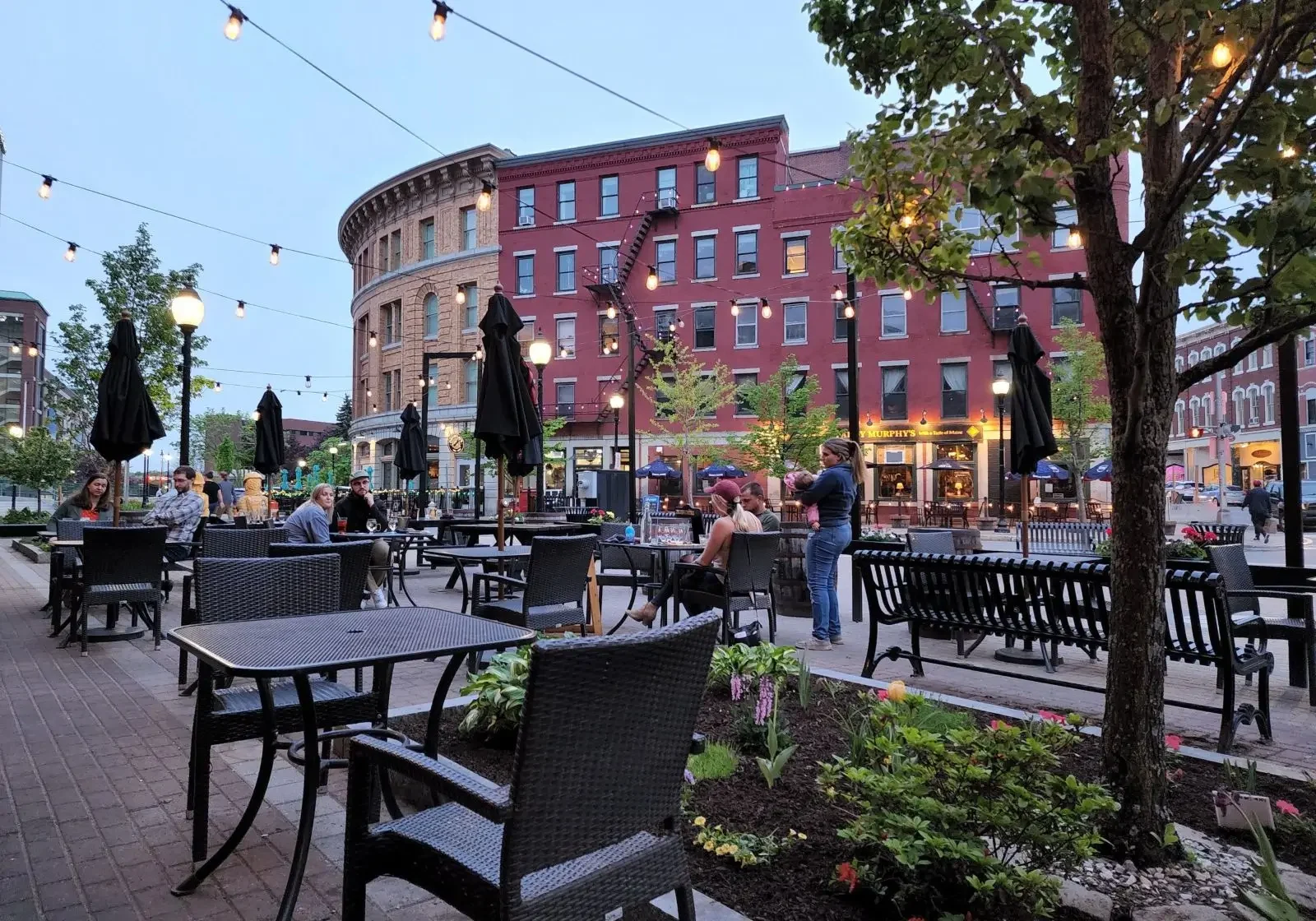 Outdoor dining area with tables, chairs, and string lights in an urban setting at dusk.